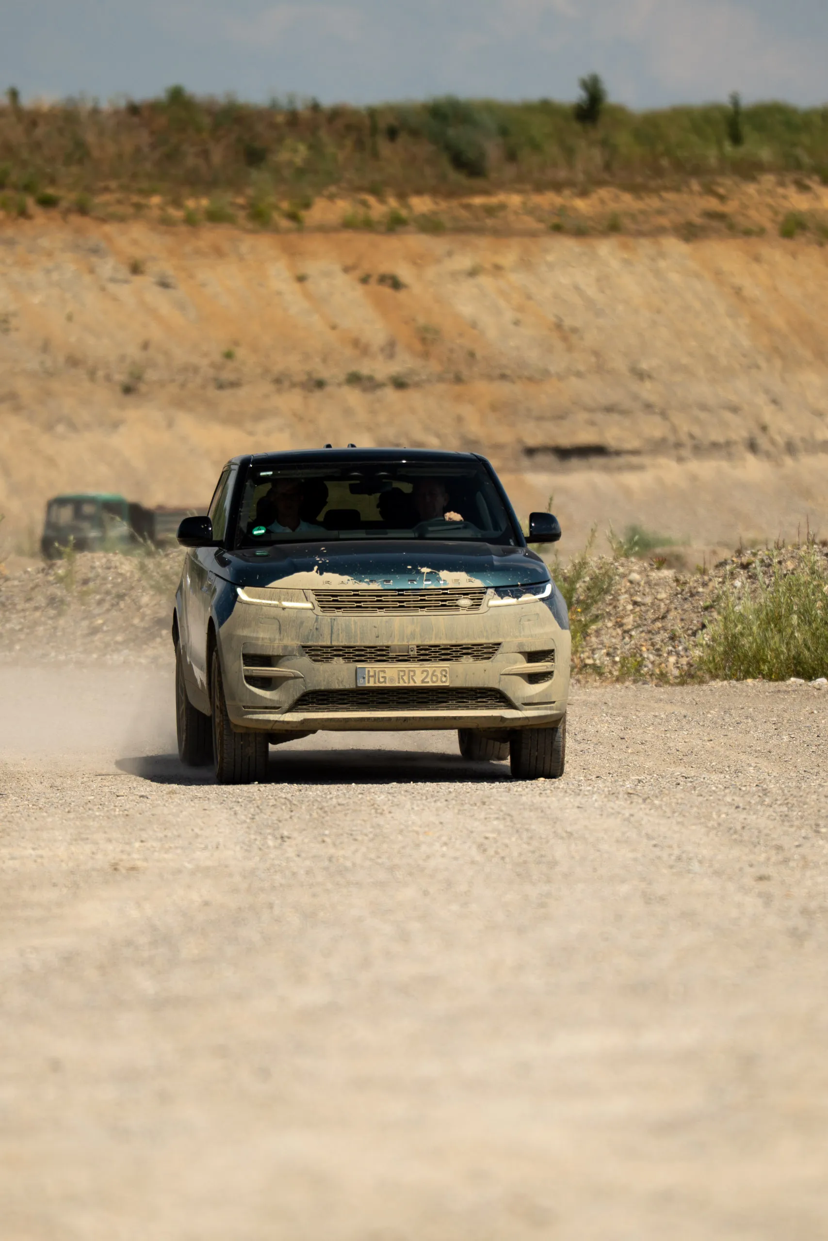 A Range Rover Sport caked in mud drives along a gravel track, the terraced wall of a quarry behind it.