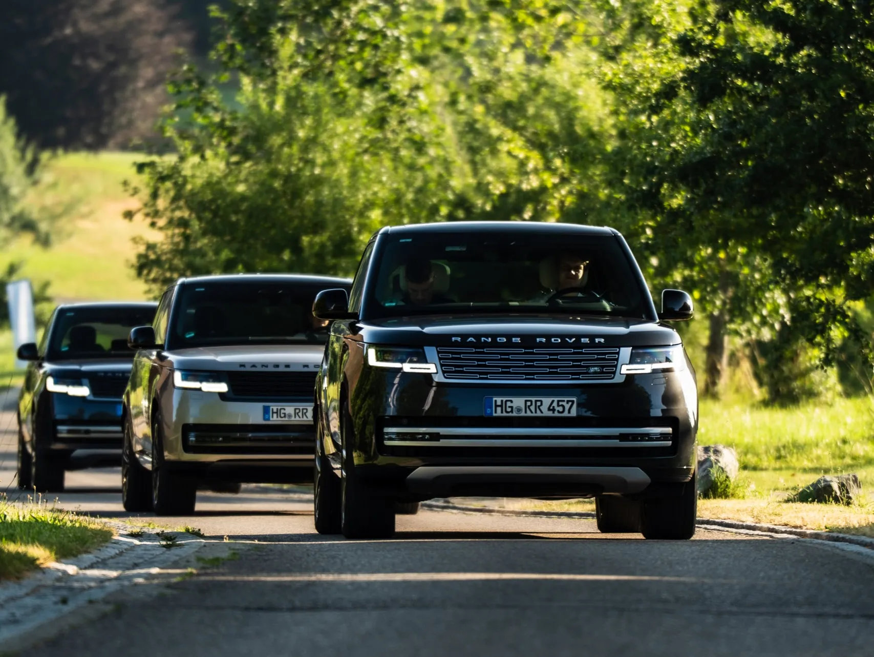 Drei Range Rover fahren hintereinander über eine schmale Landstraße zwischen Wiesen und Bäumen; im vorderen Fahrzeug sind Fahrer und Beifahrer zu erkennen.