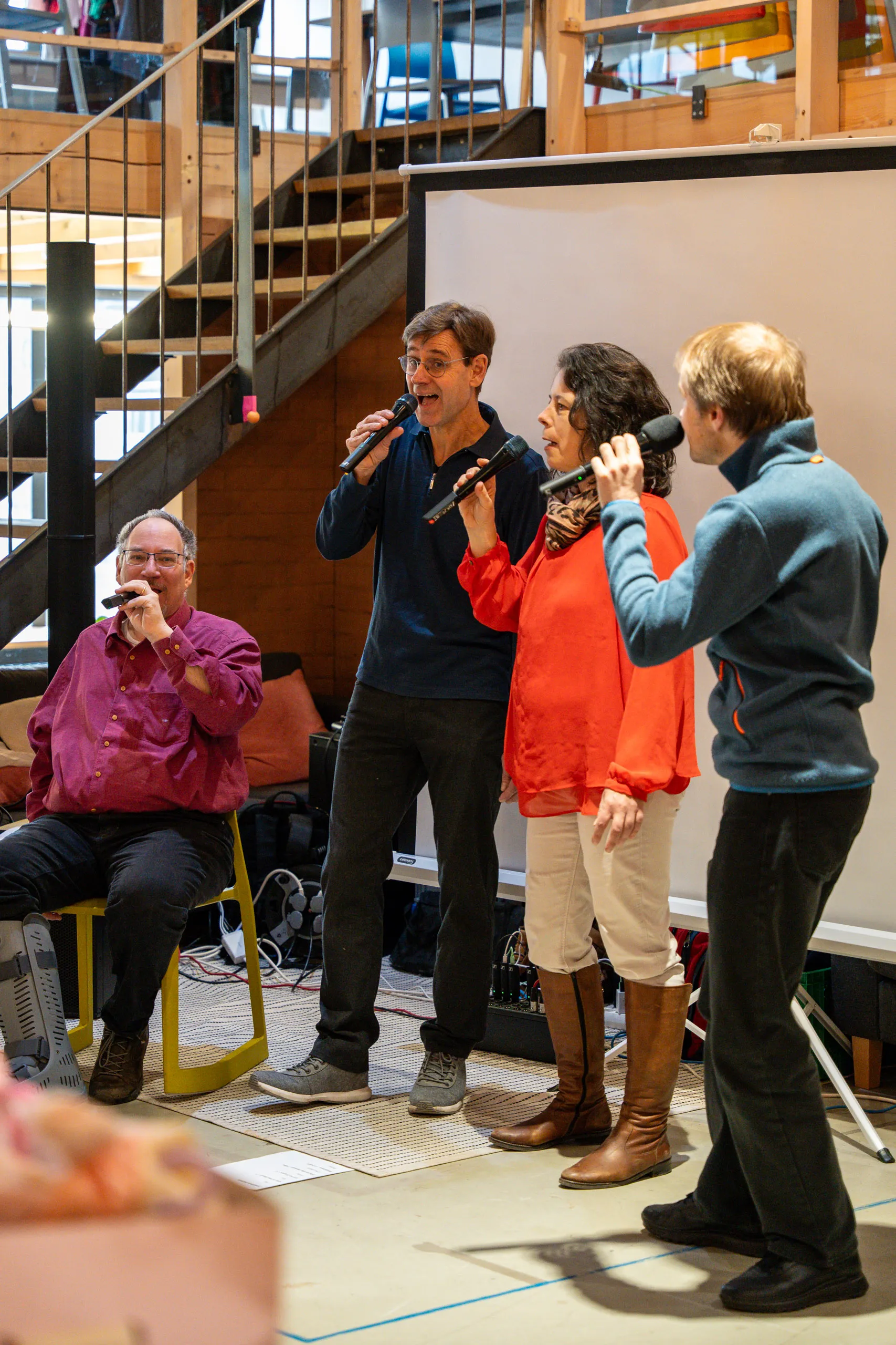 The a cappella group Sing Sang performing with microphones at the secondhand bazaar, a projection screen and the staircase behind them.