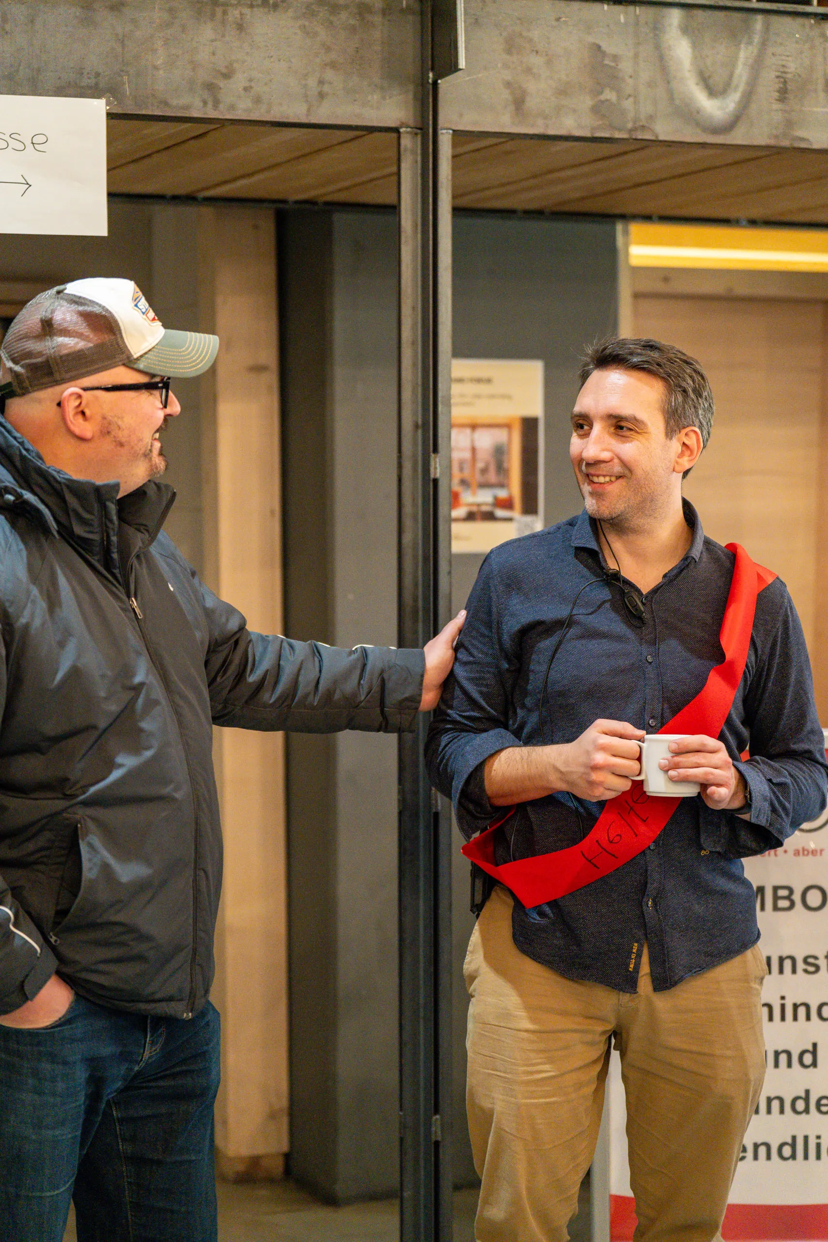 Daniel Musizza, holding a coffee cup and wearing a red sash, in conversation with a visitor at the secondhand bazaar, next to the sign pointing to the till.