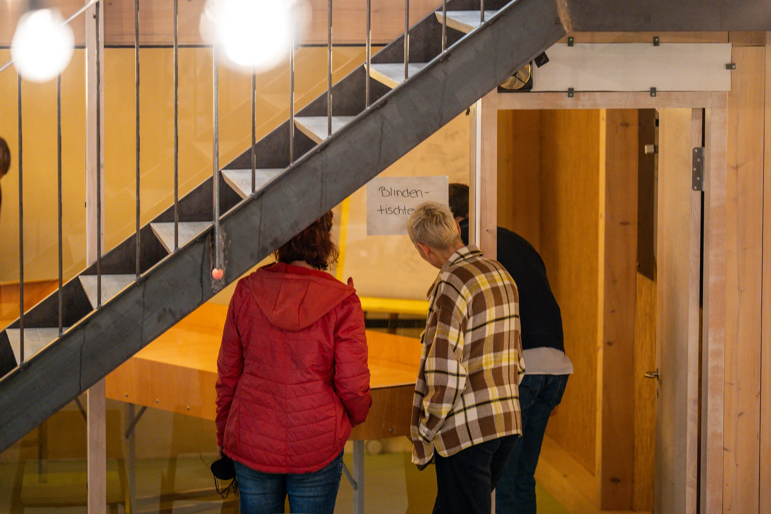 Two visitors at the blind table tennis table under the staircase, beside a handwritten sign reading "Blindentischtennis".