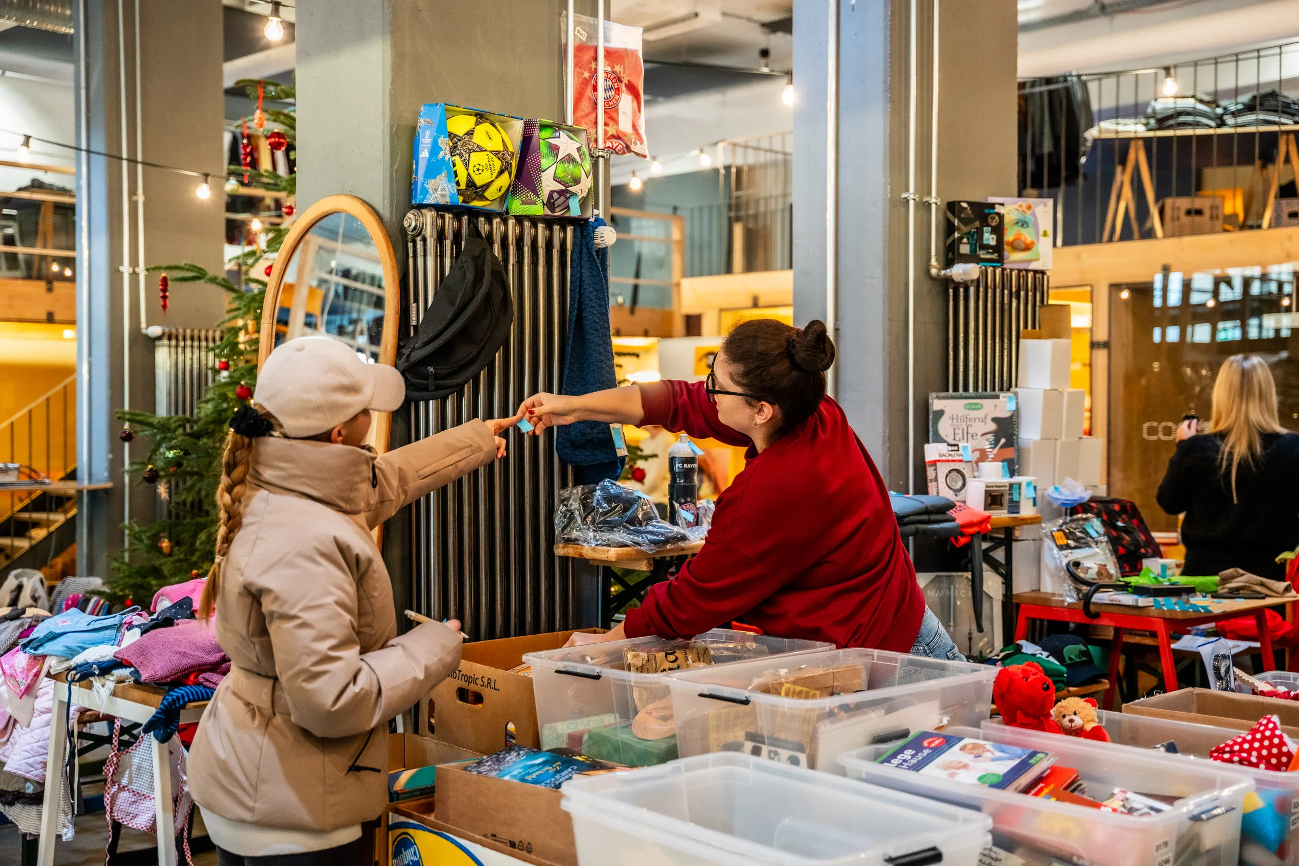 At a stall at the secondhand bazaar, a volunteer in a red jumper hands a young visitor a sales slip, with a decorated Christmas tree behind them.