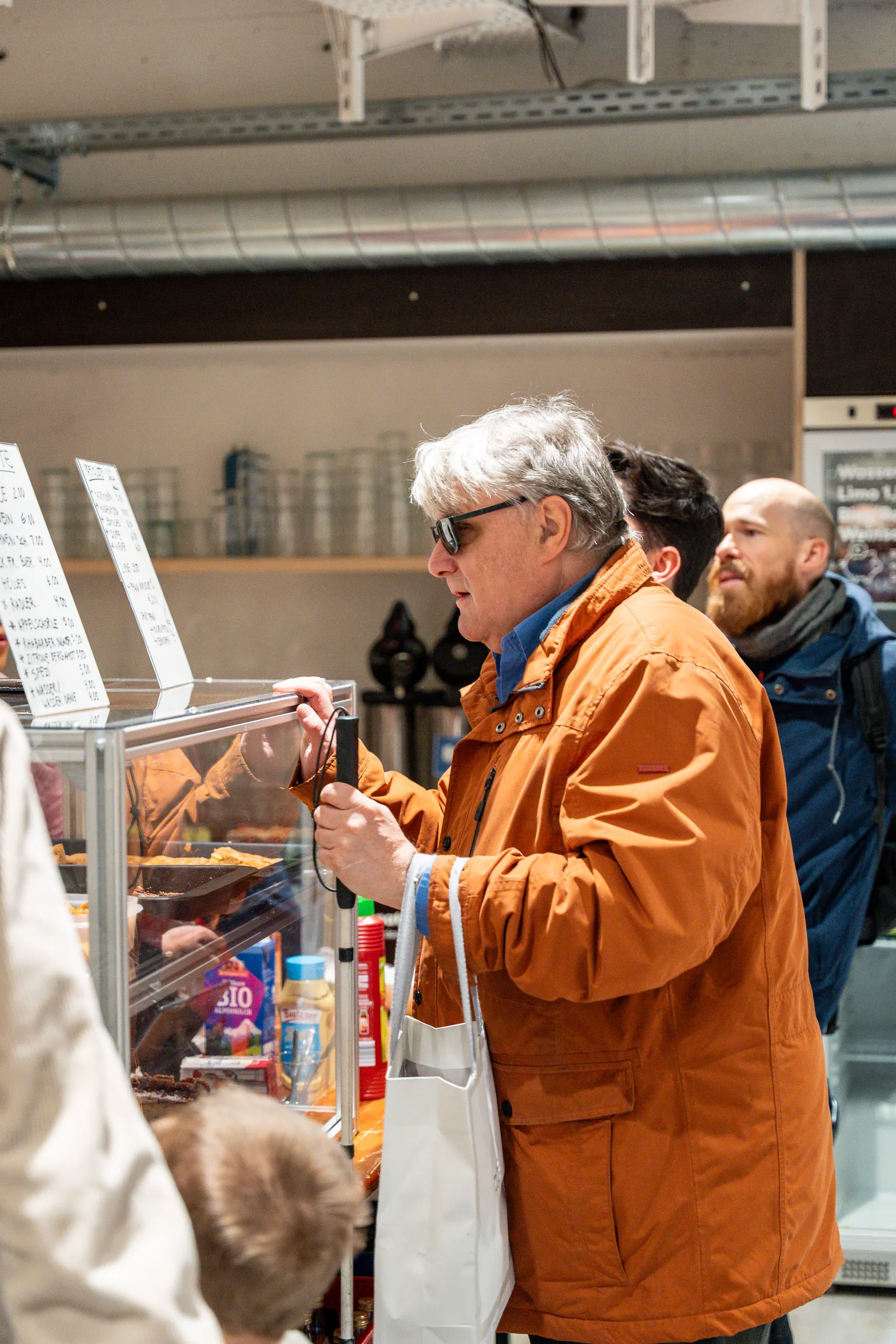 A blind visitor with a white cane and sunglasses at the food and drinks counter of the bazaar.