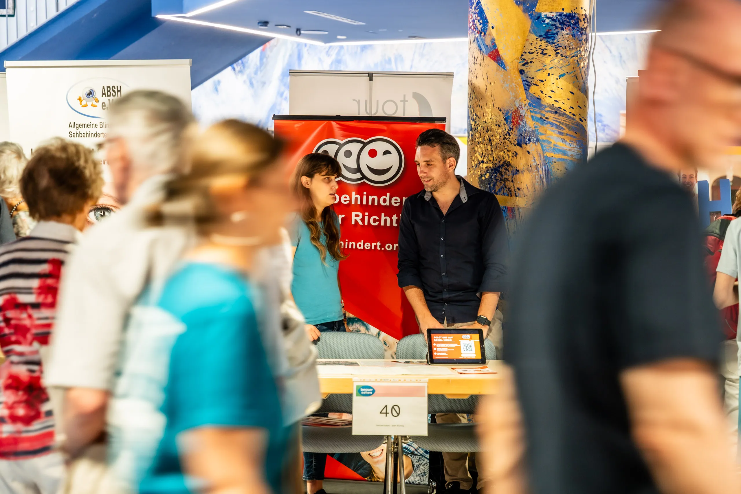 Daniel Musizza, chairman of Sehbehindert – aber Richtig e. V., and a colleague at their stand at the besser sehen trade fair, a climbing wall behind them and visitors passing in the foreground