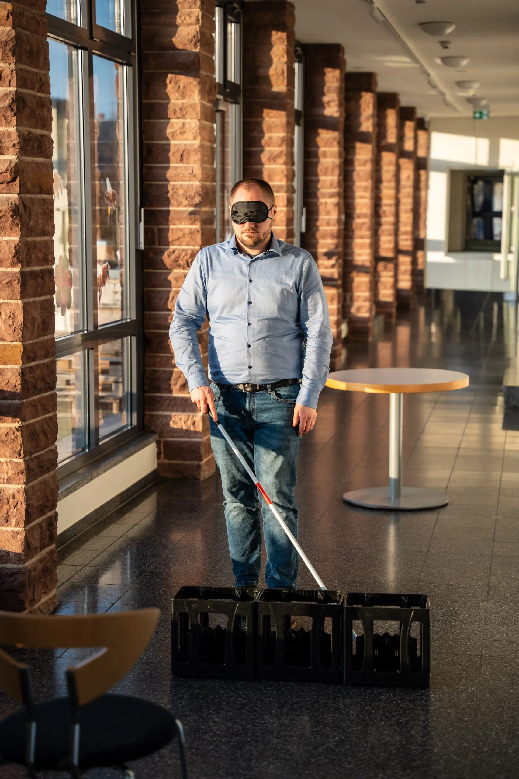 Ein Mann mit schwarzer Augenmaske ertastet sich mit dem Langstock den Weg an aufgestellten Getränkekisten vorbei, in einem sonnendurchfluteten Foyer mit Backsteinpfeilern. Übung zum Perspektivwechsel.