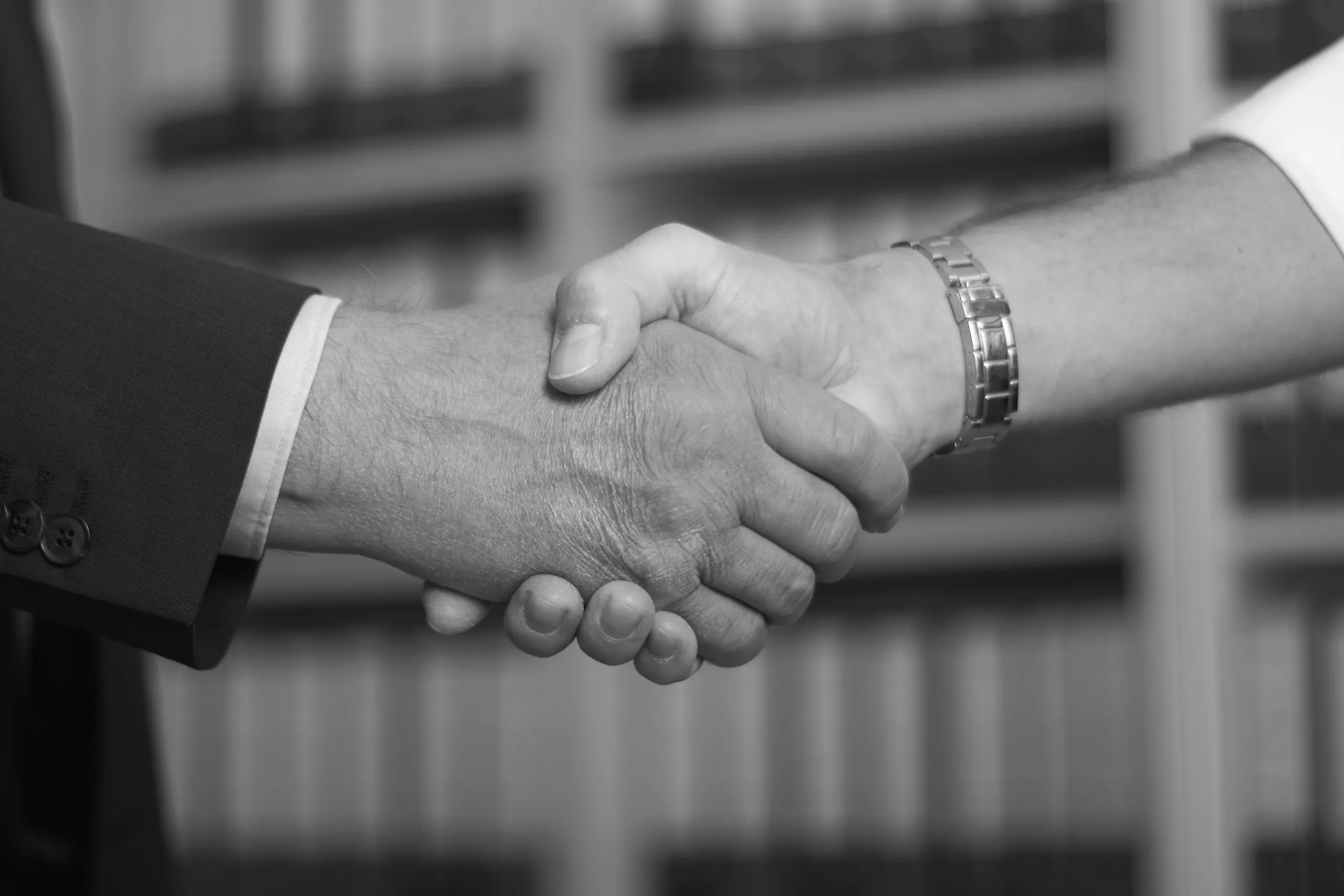 A black-and-white close-up of Ralph Steinmann and Jörg Müller shaking hands at the Steinmann Tax Consulting offices, with the filing cabinets blurred in the background.