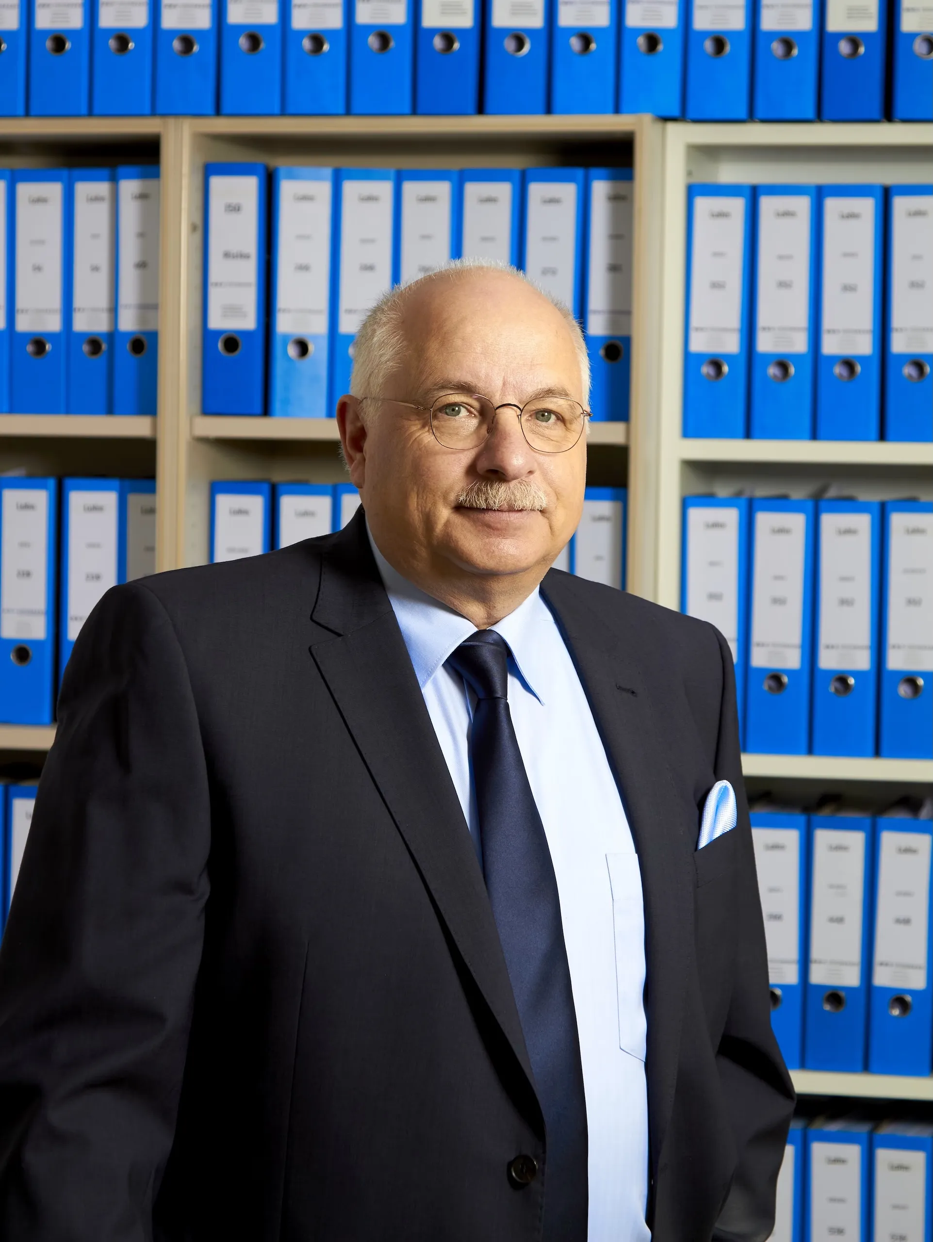 A business portrait of Ralph Steinmann in a dark suit and tie, standing in front of a shelf full of blue file folders at the offices of Steinmann Steuerberatung mbH.