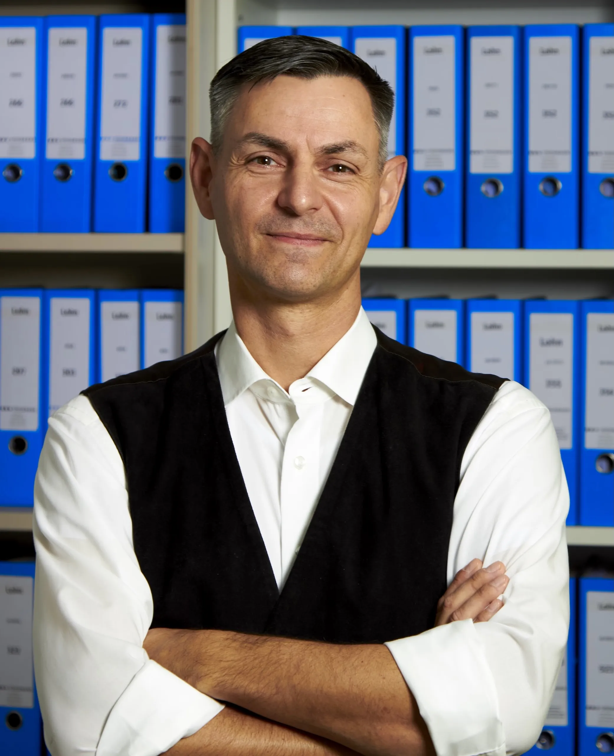 A business portrait of Jörg Müller in a white shirt and black vest, with his arms crossed in front of the blue file folders of Steinmann Steuerberatung mbH.