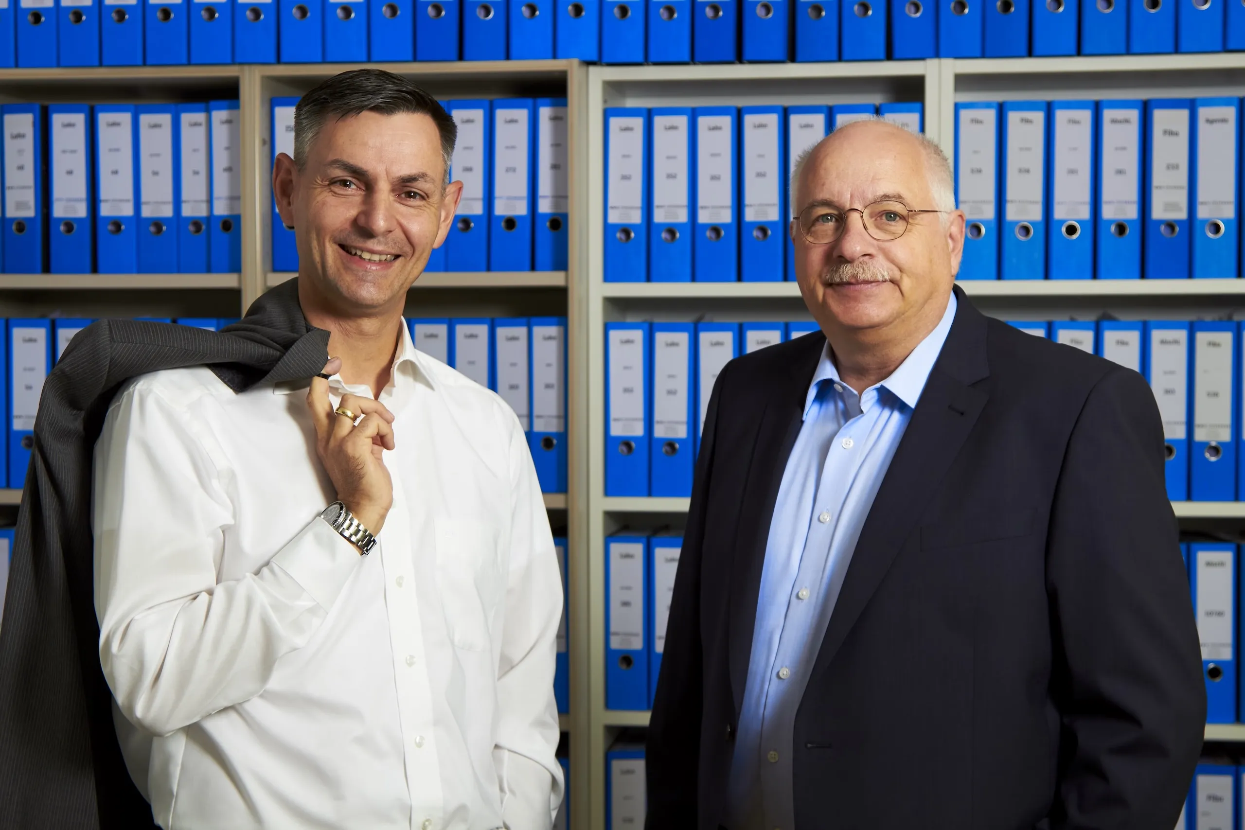 Jörg Müller und Ralph Steinmann von der Steinmann Steuerberatung nebeneinander vor einem Regal mit blauen Aktenordnern in den Kanzleiräumen.